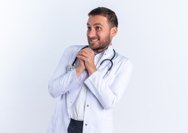 Médico jovem com jaleco branco e estetoscópio pendurado no pescoço de mãos dadas, feliz e alegre, esperando por algo
