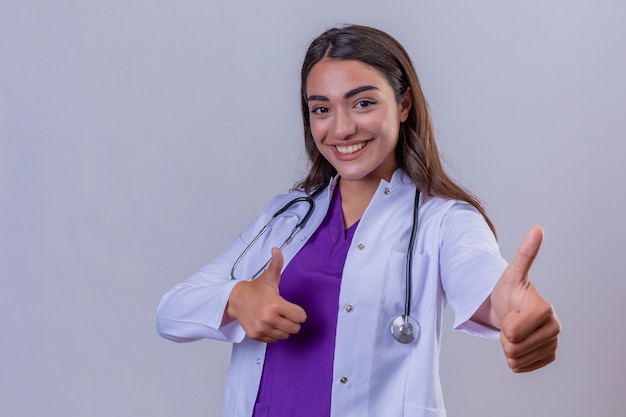 Médico jovem de jaleco branco com estetoscópio com grande sorriso mostrando sinal de ok polegares para cima, sobre fundo branco