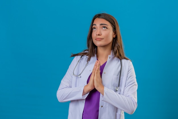 Médico jovem de jaleco branco com estetoscópio, olhando as mãos postas em gesto de oração, esperando o gesto sobre fundo azul isolado