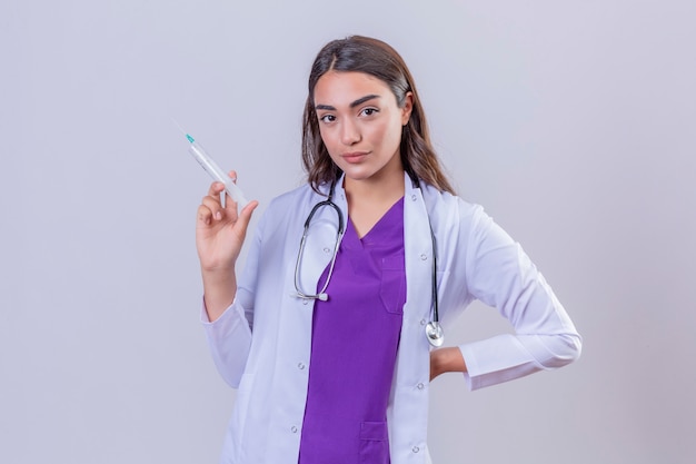 Médico jovem de jaleco branco com estetoscópio olhando confiante segurando a seringa com vacina médica sobre fundo branco isolado
