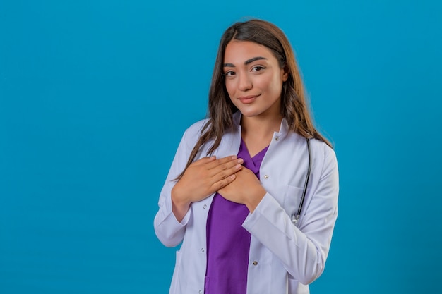 Médico jovem de jaleco branco com estetoscópio sorrindo com as mãos no peito e gesto agradecido no rosto sobre fundo azul isolado