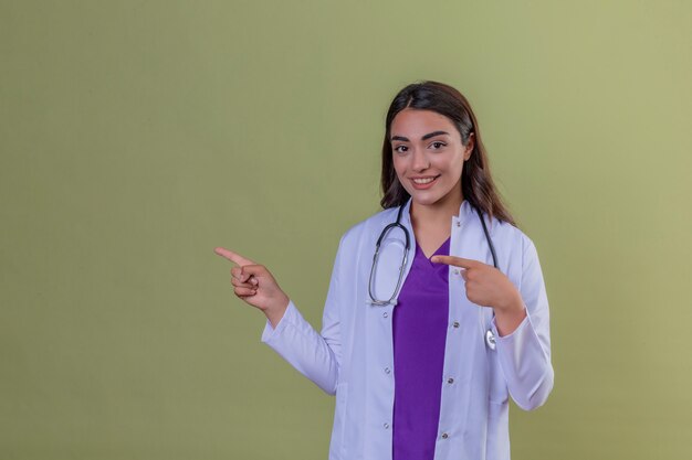 Médico jovem de jaleco branco com estetoscópio sorrindo e olhando para a câmera apontando com as duas mãos e dedos para o lado de pé sobre fundo verde isolado