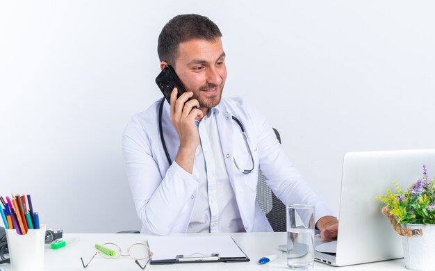 Médico jovem de jaleco branco e com estetoscópio sorrindo alegremente sentado à mesa com laptop trabalhando falando no celular em branco
