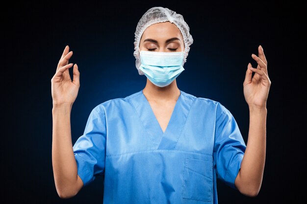 Médico jovem de uniforme azul com os olhos fechados, segurando as mãos levantadas