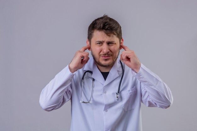 Médico jovem vestindo jaleco branco e estetoscópio cobrindo os ouvidos com os dedos com expressão irritada pelo barulho