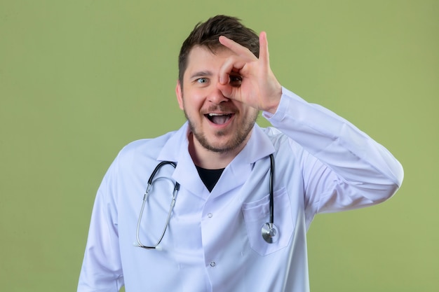 Médico jovem vestindo jaleco branco e estetoscópio, fazendo sinal de ok com a mão e os dedos olhando através de sinal com um grande sorriso sobre fundo verde isolado