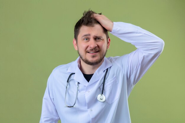 Médico jovem vestindo jaleco branco e estetoscópio surpreso com cara feliz sobre fundo verde isolado