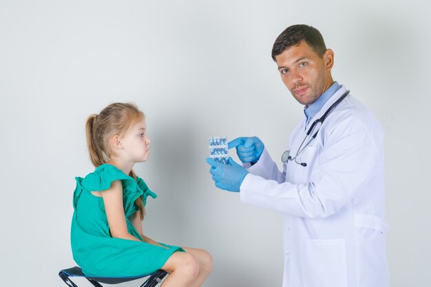 Médico mostrando comprimidos enquanto criança sentada exausta em vista frontal uniforme de branco.