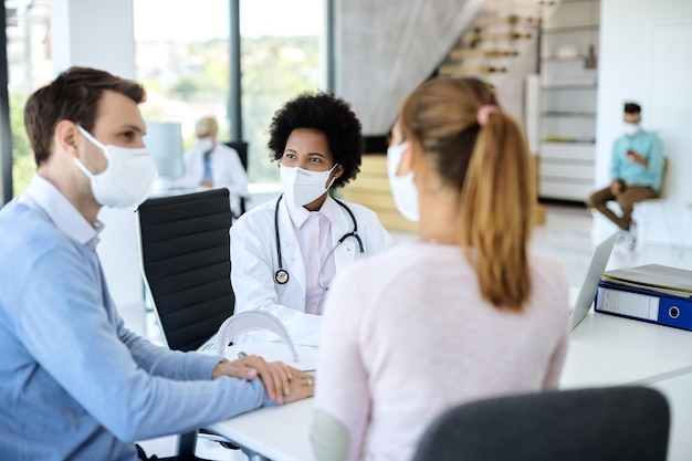 Médico negro sorridente com máscara facial conversando com um casal durante aconselhamento médico na clínica