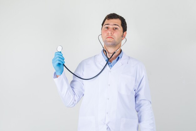 Médico olhando para a câmera enquanto segura o estetoscópio com jaleco branco, luvas e olhando sério