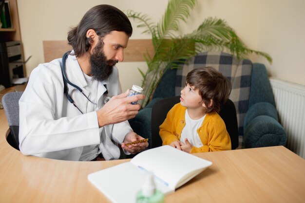 Médico pediatra que examina uma criança em consultório médico comfortabe. Conceito de saúde, infância, medicina, proteção e prevenção. Garotinho confia no médico e sente emoções calmas e positivas.