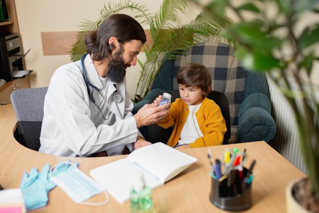 Médico pediatra que examina uma criança em consultório médico comfortabe. Conceito de saúde, infância, medicina, proteção e prevenção. Garotinho confia no médico e sente emoções calmas e positivas.