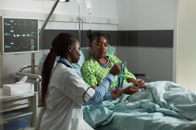 Médico praticante afro-americano ao lado de um paciente jovem doente durante a consulta médica explicando a experiência em raios X de ossos na enfermaria do hospital. Mulher médica discutindo conhecimentos de saúde