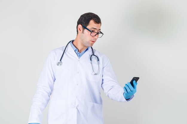Médico segurando e olhando para smartphone em vista frontal de óculos, jaleco branco, luvas.