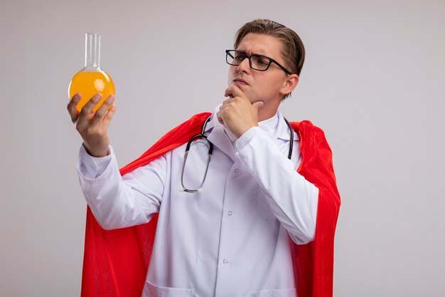 Médico super-herói homem vestindo jaleco branco com capa vermelha e óculos com estetoscópio em volta do pescoço segurando um frasco com um líquido amarelo olhando para ele perplexo em pé sobre uma parede branca