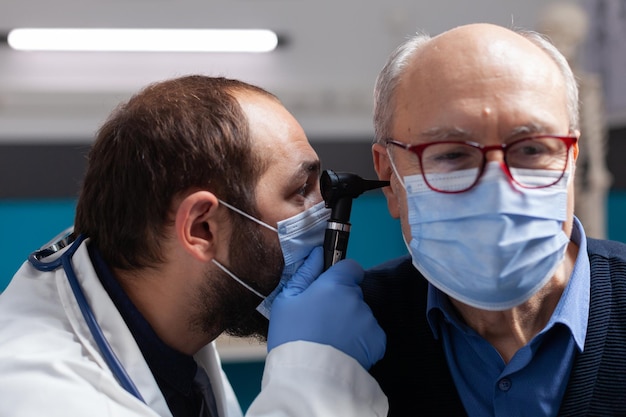 Médico usando otoscópio para fazer exame de ouvido em homem sênior, fazendo consulta de otorrinolaringologia com o paciente. Médico com máscara facial, examinando a infecção para administrar o tratamento prescrito. Fechar-se