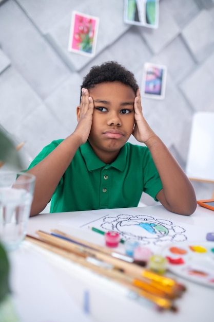 Meditação. Menino de pele escura pensativo em idade escolar primária em camiseta verde tocando a cabeça com as mãos olhando de lado sentado à mesa com desenho e pincéis à luz do dia
