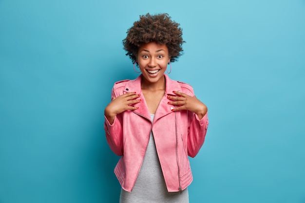 Meia foto de uma garota afro-americana bem alegre vestida com uma jaqueta rosa da moda, sorrindo amplamente, ouve boas notícias emocionantes, poses