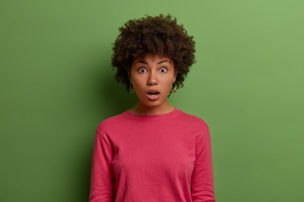 Foto grátis meia foto de uma mulher envergonhada e muda de cabelo com cabelo afro, olhos esbugalhados de espanto, abre a boca em surpresa, reage a notícias surpreendentes, vestida com um macacão rosa, isolada na parede verde