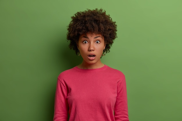 Meia foto de uma mulher envergonhada e muda de cabelo com cabelo afro, olhos esbugalhados de espanto, abre a boca em surpresa, reage a notícias surpreendentes, vestida com um macacão rosa, isolada na parede verde