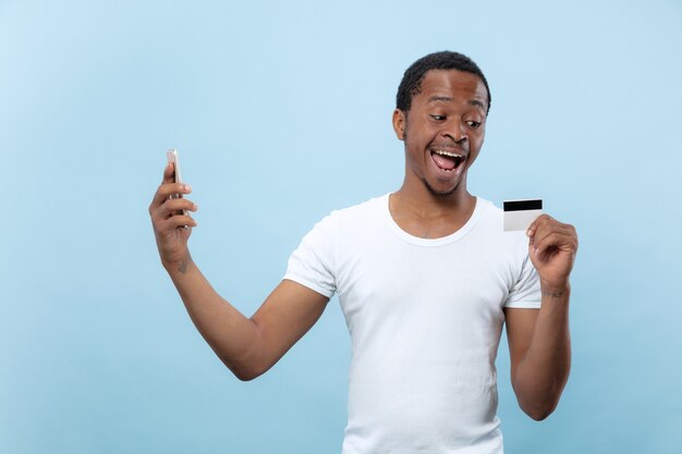 Meia retrato de um jovem afro-americano de camisa branca, segurando um cartão e um smartphone sobre fundo azul. Emoções humanas, expressão facial, anúncio, vendas, finanças, conceito de pagamentos online.