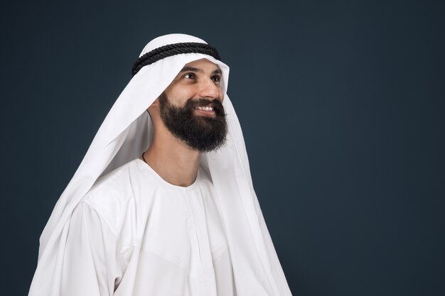 Meia retrato do empresário Arábia Saudita na parede azul escura. Jovem modelo masculino em pé e sorrindo. Conceito de negócios, finanças, expressão facial, emoções humanas.