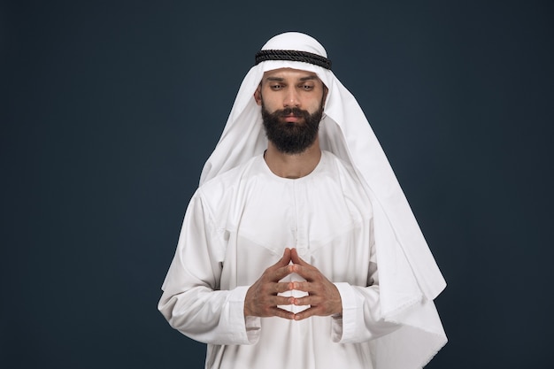 Meia retrato do empresário da arábia saudita em fundo azul escuro do estúdio. jovem modelo masculino orando e parece pensativo. conceito de negócios, finanças, expressão facial, emoções humanas.