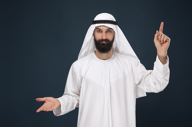 Meia retrato do homem saudita árabe em fundo azul escuro do estúdio. jovem modelo masculino sorrindo e apontando. conceito de negócios, finanças, expressão facial, emoções humanas, tecnologias.