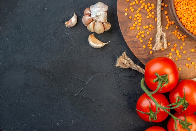 Meia vista de lentilha vermelha em alhos de corda de tigela marrom em tomates de tábua redonda de madeira com hastes em fundo preto