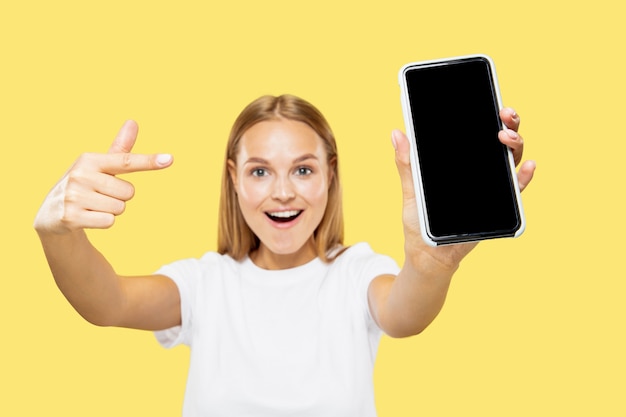 Meio comprimento do retrato de mulher jovem caucasiana em fundo amarelo do estúdio. bela modelo feminino em camisa branca. conceito de emoções, expressão facial, vendas, pagamento online. mostrando a tela do telefone.