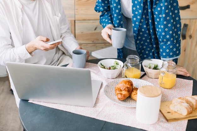 Meio de homens segurando a xícara de café perto delicioso café da manhã com suco e laptop sobre a mesa de madeira