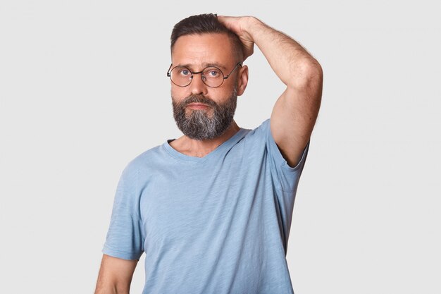 Meio envelhecido homem barbudo bonito com óculos elegantes vestindo luz azul camiseta casual. modelo de trabalho duro coloca isolado na luz.