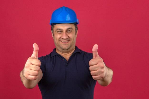 Meio envelhecido homem vestindo camisa polo e capacete de segurança com sorriso no rosto e aparecendo os polegares sobre parede rosa isolada