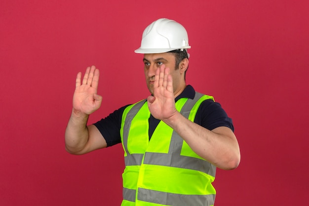 Meio envelhecido homem vestindo construção colete amarelo e capacete de segurança, guiando alguém para a direção parar gesto sobre parede rosa isolada