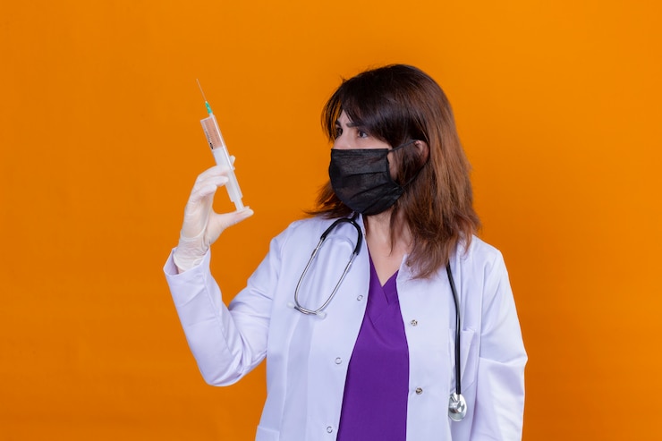 Foto grátis meio envelhecido médico vestindo jaleco branco na máscara facial protetora preta e com estetoscópio segurando a seringa olhando para ele