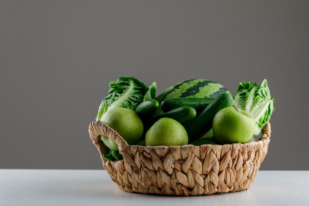 Melancia com alface, maçã, pepino em uma cesta de vime na mesa branca e cinza