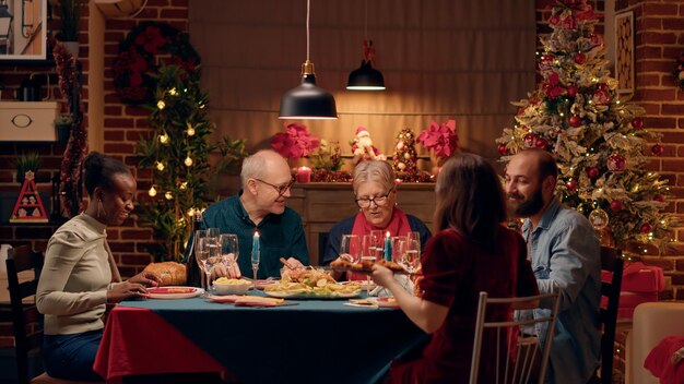 Membros da família multicultural positiva comemorando o Natal com jantar especial em casa. Pessoas felizes desfrutando de comida caseira tradicional enquanto se divertem juntos. Tiro de tripé