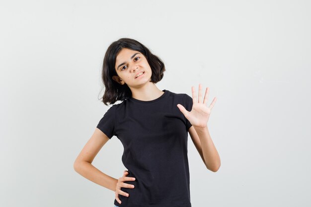 Menina acenando com a mão para dizer Olá, em t-shirt preta, vista frontal.