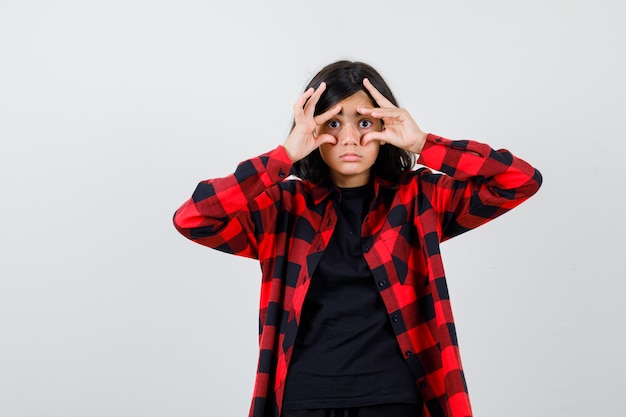 Menina adolescente abrindo os olhos com os dedos na camiseta, camisa quadriculada e olhando com foco, vista frontal.