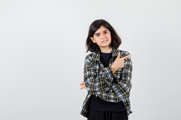 Menina adolescente apontando para o canto superior direito em uma camisa casual e olhando alegre, vista frontal.