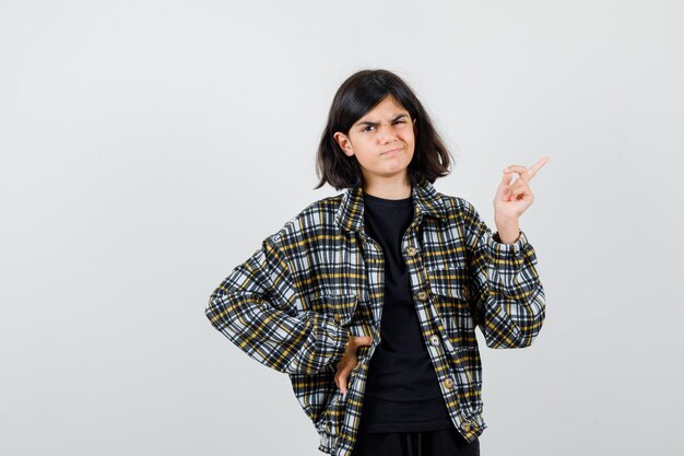 Menina adolescente apontando para o canto superior direito, segurando a mão na cintura em uma camisa casual e parecendo desapontada. vista frontal.