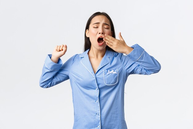 Menina adolescente asiática com sono, de pijama, bocejando e se espreguiçando, acordando cedo do alarme. Mulher fofa cansada em pijamas em pé, fundo branco querendo dormir