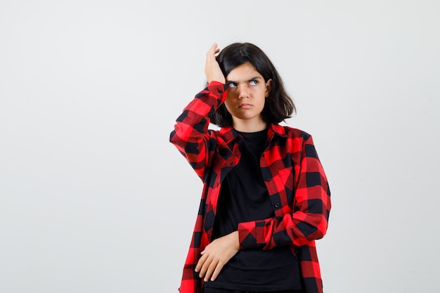Menina adolescente coçando a cabeça enquanto olha para o lado em t-shirt, camisa xadrez e olhando com raiva, vista frontal.
