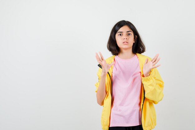 Menina adolescente com agasalho amarelo, t-shirt espalhando as palmas das mãos e parecendo chocada, vista frontal.