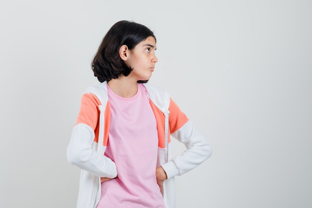 Menina adolescente com jaqueta, camisa rosa, olhando para longe, segurando a mão na cintura e olhando com foco.