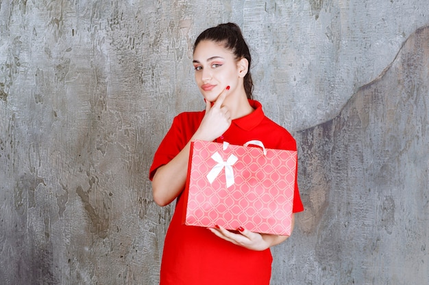 Menina adolescente de camisa vermelha, segurando uma sacola de compras vermelha e parece confusa e pensativa.