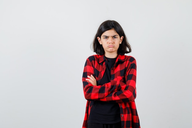Foto grátis menina adolescente em pé com os braços cruzados em t-shirt, camisa xadrez e olhando ressentida, vista frontal.