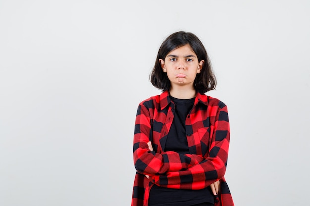 Menina adolescente em pé com os braços cruzados em t-shirt, camisa xadrez e olhando ressentida, vista frontal.