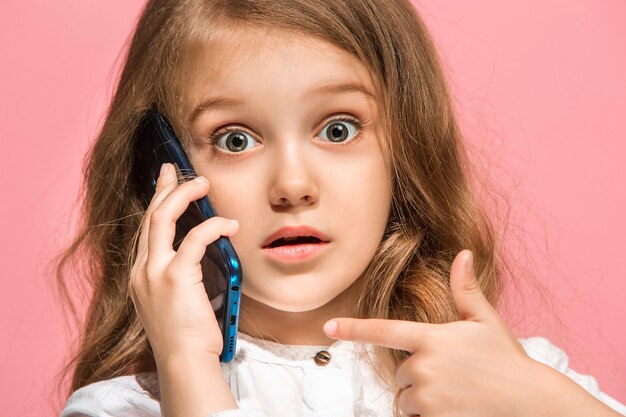 Menina adolescente em pé, sorrindo com o celular sobre o fundo rosa da moda do estúdio.