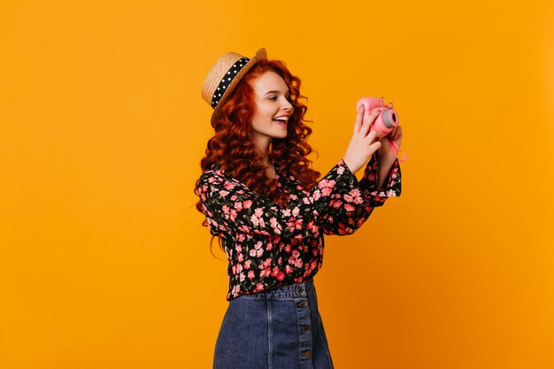 Menina adolescente em saia jeans e blusa elegante feliz faz foto, segurando a mini câmera no espaço laranja.